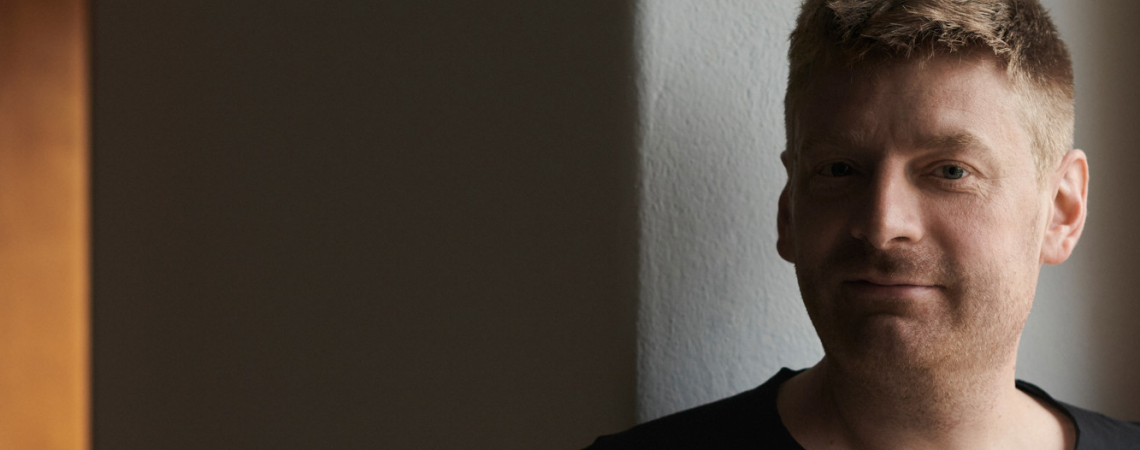 Mann in schwarzem T-Shirt steht vor einer dunklen, schlichten Wand.