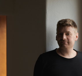 Mann in schwarzem T-Shirt steht vor einer dunklen, schlichten Wand.