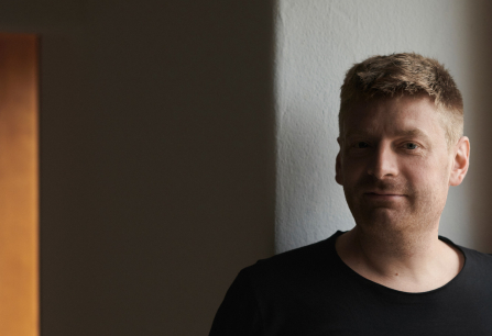 Mann in schwarzem T-Shirt steht vor einer dunklen, schlichten Wand.