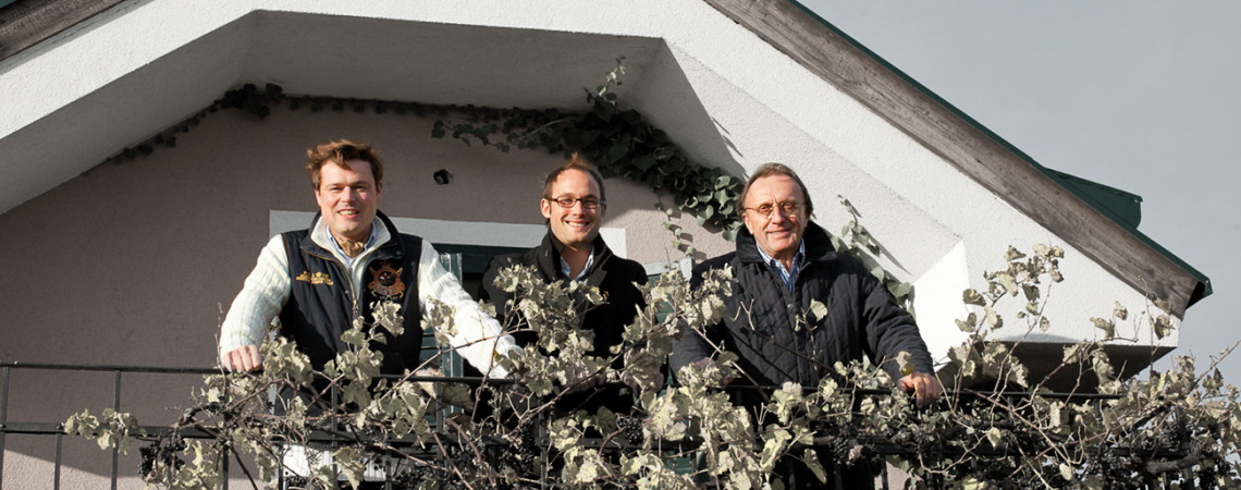 Drei Personen stehen auf einem Balkon mit Pflanzen und hellem Giebel im Hintergrund.