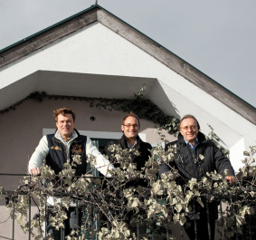 Drei Personen stehen auf einem Balkon mit Pflanzen und hellem Giebel im Hintergrund.