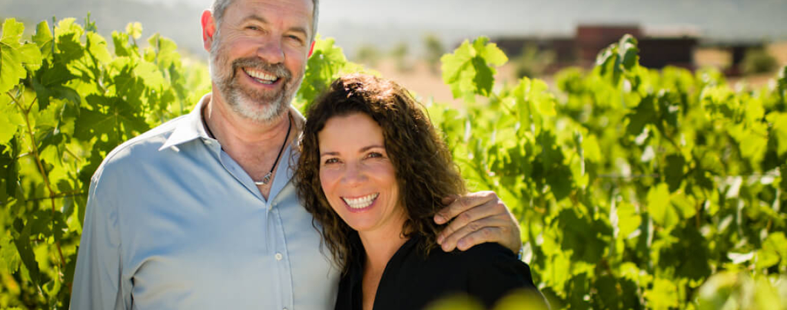Lächelndes Paar in sonnigem Weinberg vor Bergkulisse.
