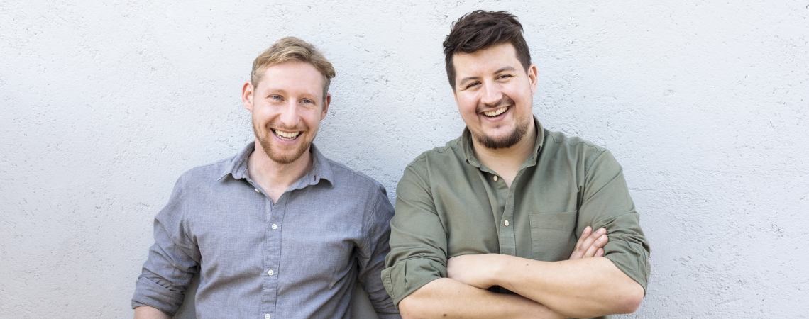Zwei lächelnde Männer vor einer hellgrauen Wand.