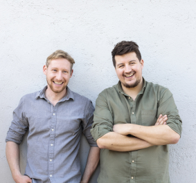 Zwei lächelnde Männer vor einer hellgrauen Wand.