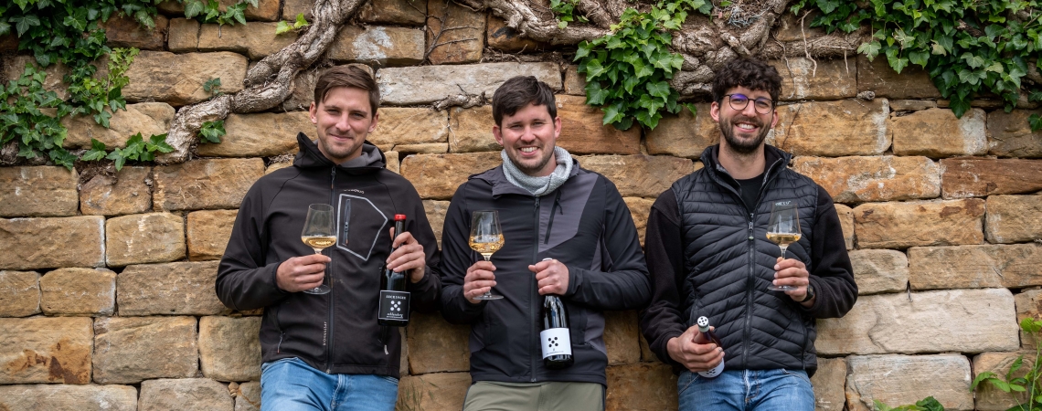 Drei Männer mit Weingläsern und Flaschen vor einer bewachsenen Steinmauer.