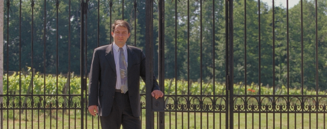 Mann im Anzug lehnt an einem geschlossenen, dekorativen Metalltor im Freien.