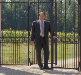 Mann im Anzug lehnt an einem geschlossenen, dekorativen Metalltor im Freien.