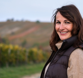 Frau lächelt auf einem Weinberg im Herbst, trägt braune Weste und Pullover.