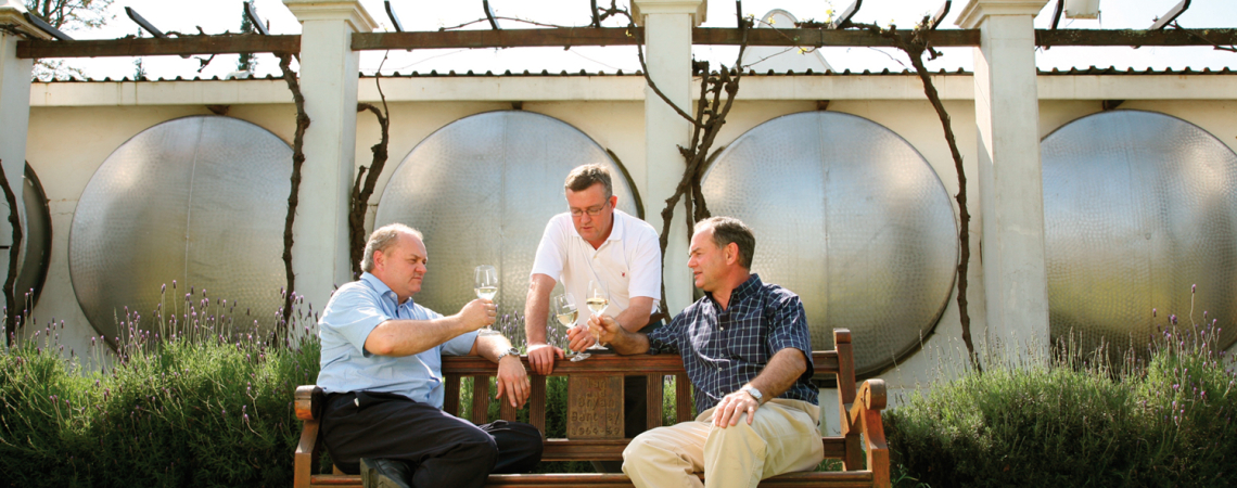 Drei Männer auf einer Holzbank, trinken Wein vor Metalltanks und Reben.