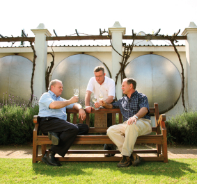 Drei Männer auf einer Holzbank, trinken Wein vor Metalltanks und Reben.