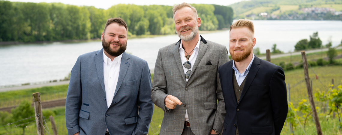 Drei Männer in Anzügen stehen lächelnd vor einem Fluss- und Wiesenhintergrund.