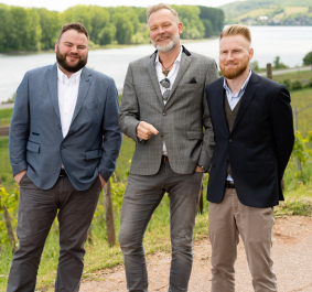 Drei Männer in Anzügen stehen lächelnd vor einem Fluss- und Wiesenhintergrund.
