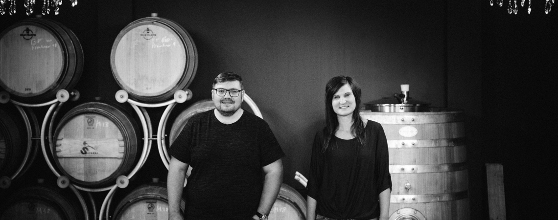 Zwei Personen vor Weinfässern und Kronleuchtern in einem Weinkeller.