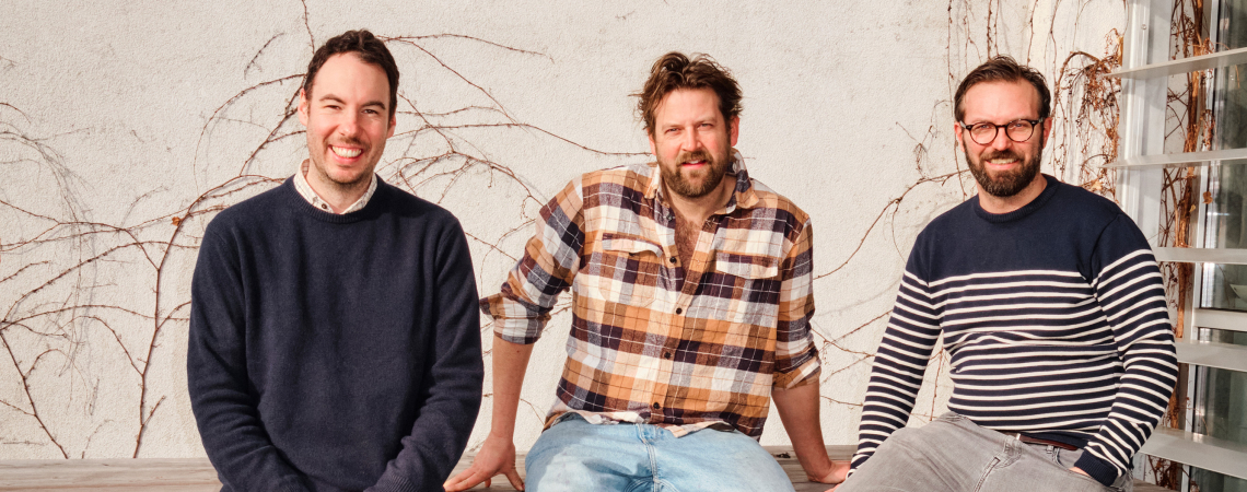 Drei lächelnde Männer sitzen entspannt vor einer mit Efeu bedeckten Wand.