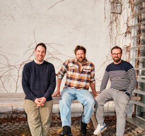 Drei lächelnde Männer sitzen entspannt vor einer mit Efeu bedeckten Wand.