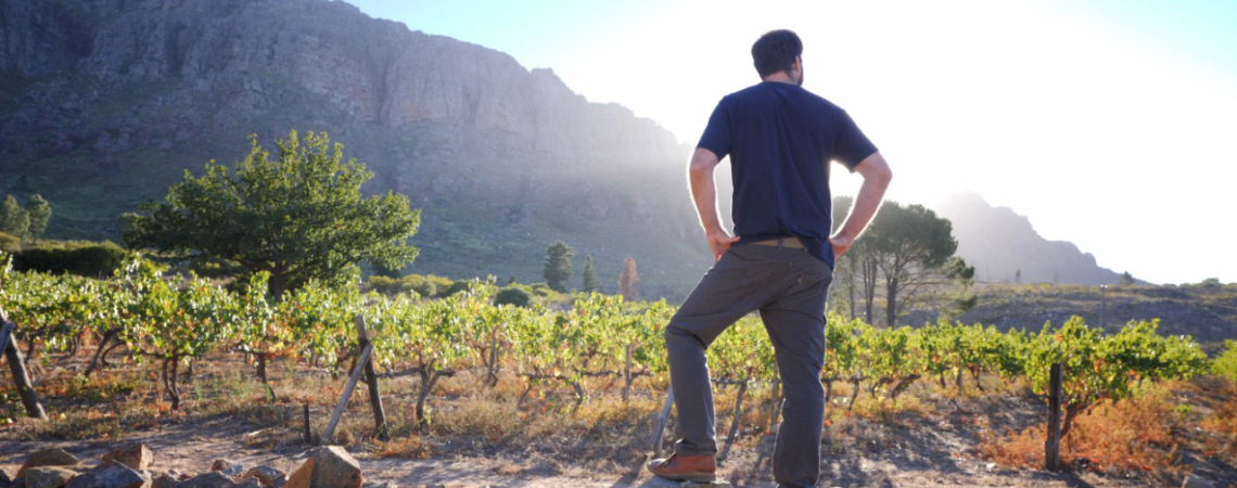 Mann steht auf Steinmauer vor Weinberg mit Bergkulisse im Sonnenlicht.