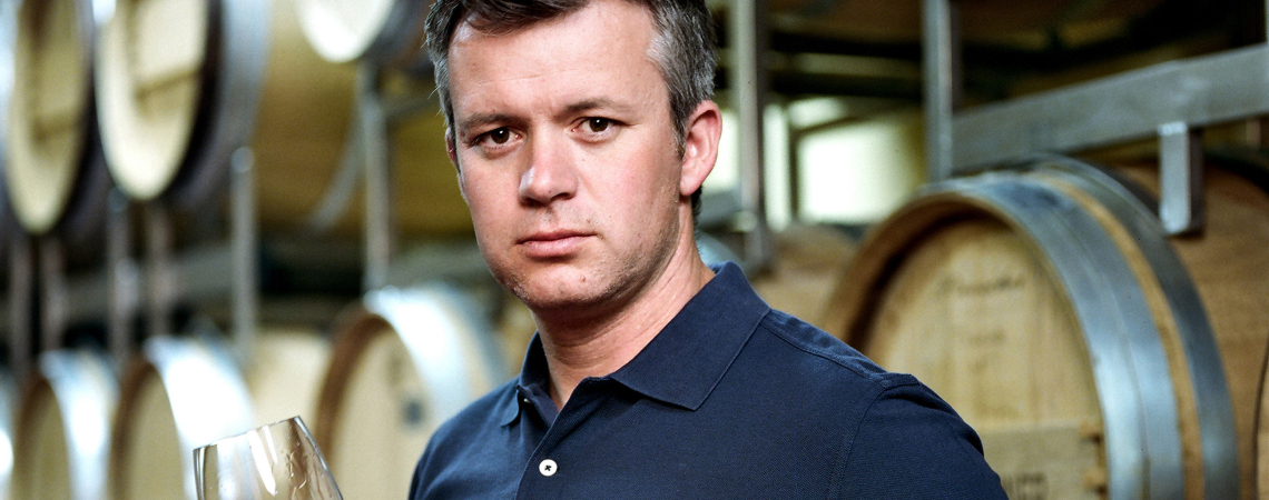 Mann in blauer Polohemd, hält Weinglas, vor Weinfässern stehend.