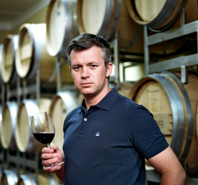 Mann in blauer Polohemd, hält Weinglas, vor Weinfässern stehend.