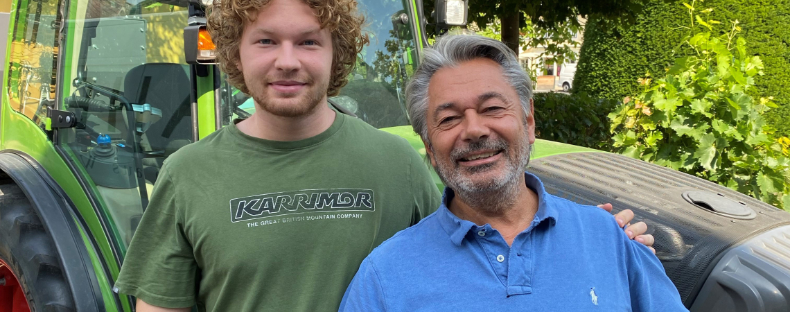 Zwei Männer lächeln vor einem grünen Traktor und üppigem Laubwerk.