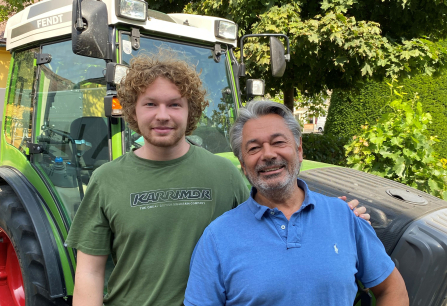 Zwei Männer lächeln vor einem grünen Traktor und üppigem Laubwerk.