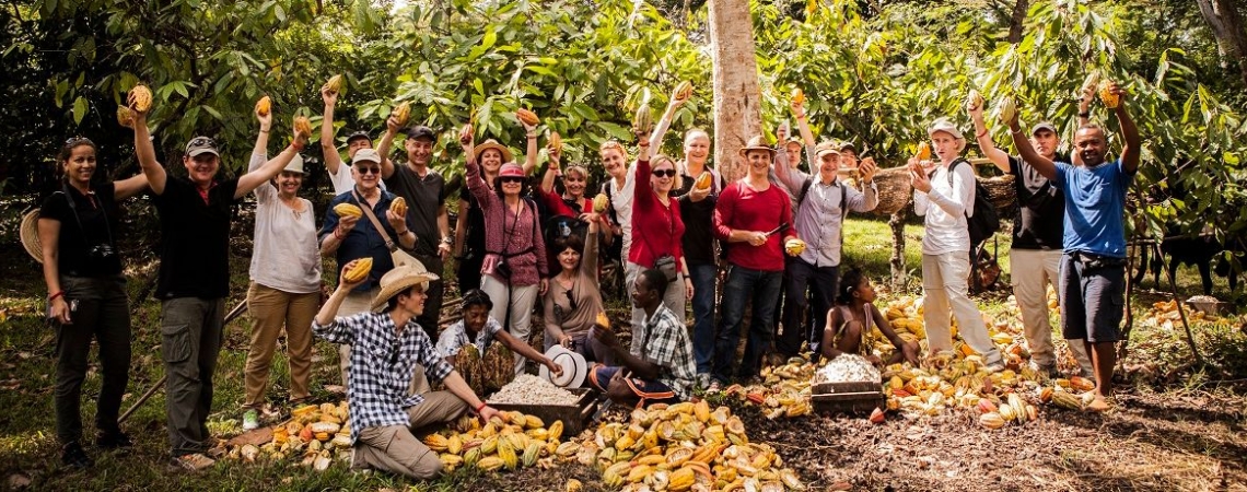 Gruppe in Plantage, lächelnd, hält frisch geerntete Früchte, umgeben von Bäumen.