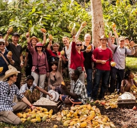 Gruppe in Plantage, lächelnd, hält frisch geerntete Früchte, umgeben von Bäumen.