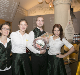 Vier Angestellte in traditioneller, eleganter Kleidung posieren lächelnd in einem Feinkostladen.