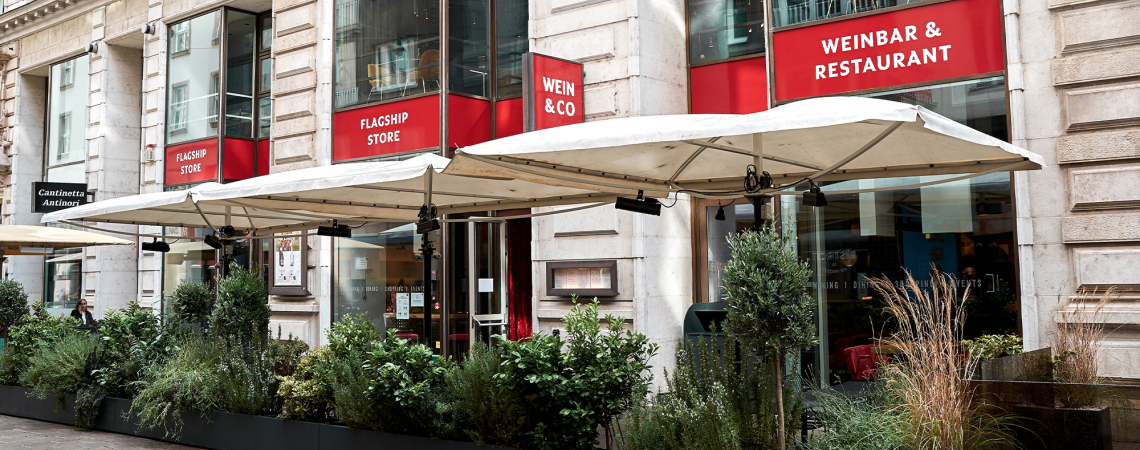 Fassade eines Restaurants mit roten Schildern und großen Sonnenschirmen davor.