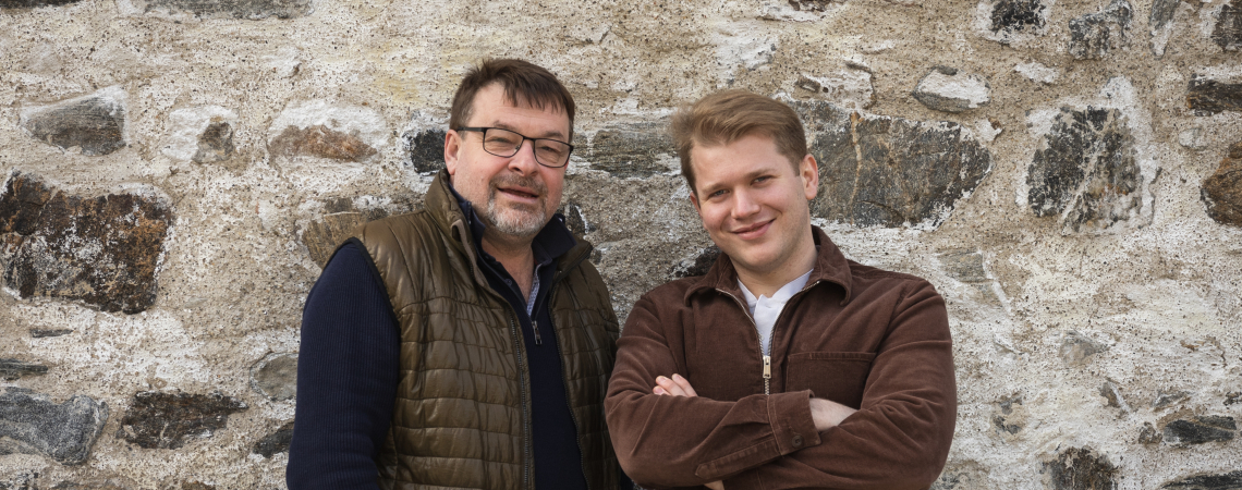 Zwei Männer vor einer rustikalen Steinmauer, lässig gekleidet und entspannt posierend.