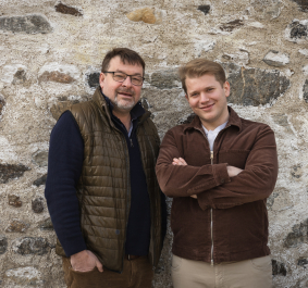 Zwei Männer vor einer rustikalen Steinmauer, lässig gekleidet und entspannt posierend.