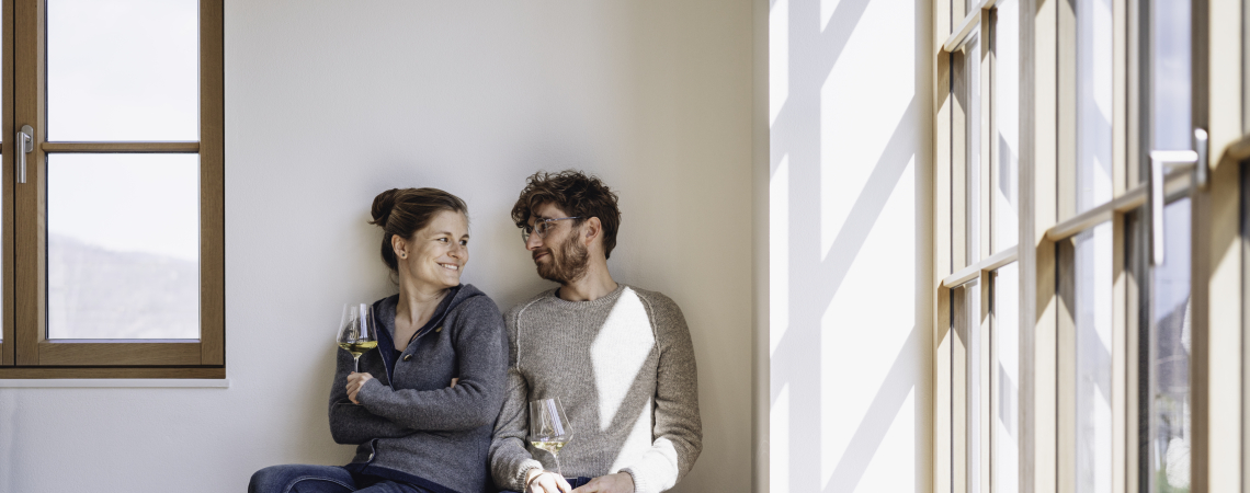 Zwei Personen sitzen lächelnd auf Holzbank, halten Weingläser, Tageslicht fällt durch Fenster.