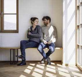 Zwei Personen sitzen lächelnd auf Holzbank, halten Weingläser, Tageslicht fällt durch Fenster.