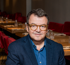 Mann in blauem Blazer sitzt in stilvollem Restaurant mit roten Stühlen.