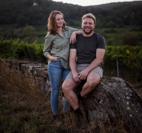 Zwei Personen lächeln auf einer alten Steinmauer in einer Weinberglandschaft.