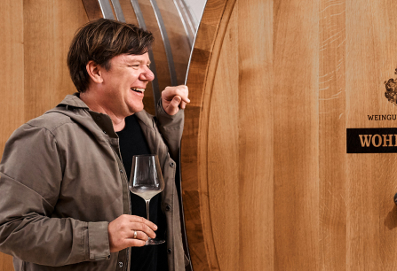 Mann lacht mit Weinglas vor großen, hölzernen Weinfässern.