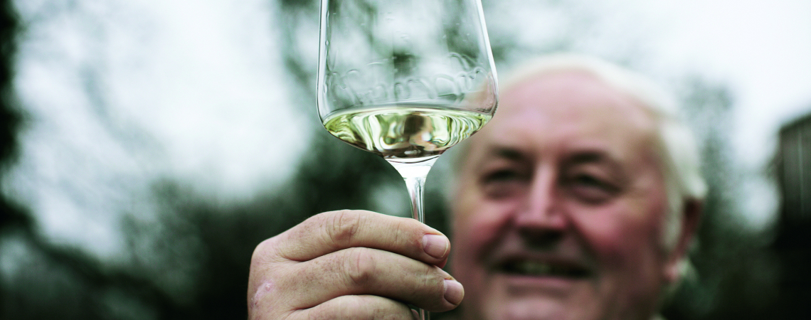 Mann hält Weinglas mit weißem Wein vor unscharfem Hintergrund.