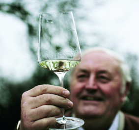 Mann hält Weinglas mit weißem Wein vor unscharfem Hintergrund.