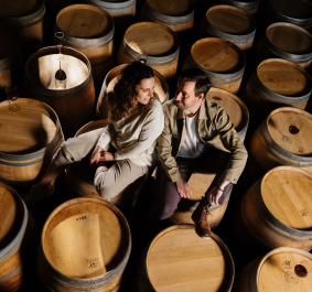 Paar sitzt auf Weinfässern in rustikalem Weinkeller, mit Weingläsern darauf.