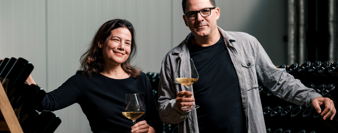 Zwei Personen mit Weingläsern posieren vor Flaschenregalen in einem Weinkeller.