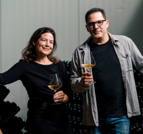 Zwei Personen mit Weingläsern posieren vor Flaschenregalen in einem Weinkeller.