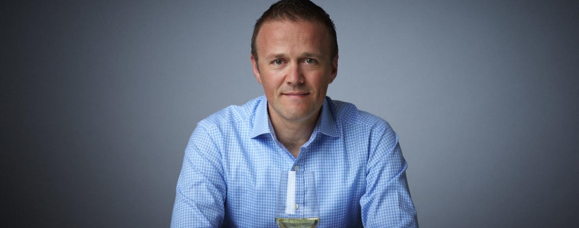 Mann in blauem Hemd mit Weinglas vor neutralem Hintergrund.