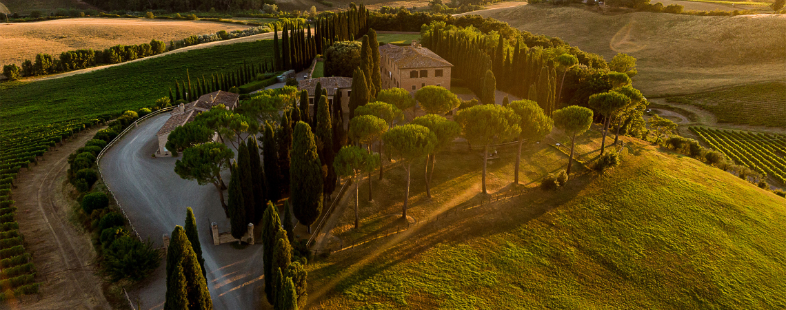 Luftaufnahme einer toskanischen Landschaft mit Villa, Zypressen und sanften Hügeln bei Sonnenuntergang.