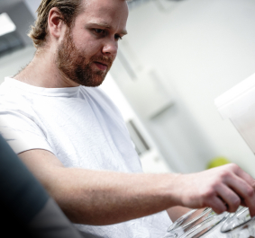 Mann in weißem T-Shirt arbeitet konzentriert mit Gläsern in einem Laborumfeld.