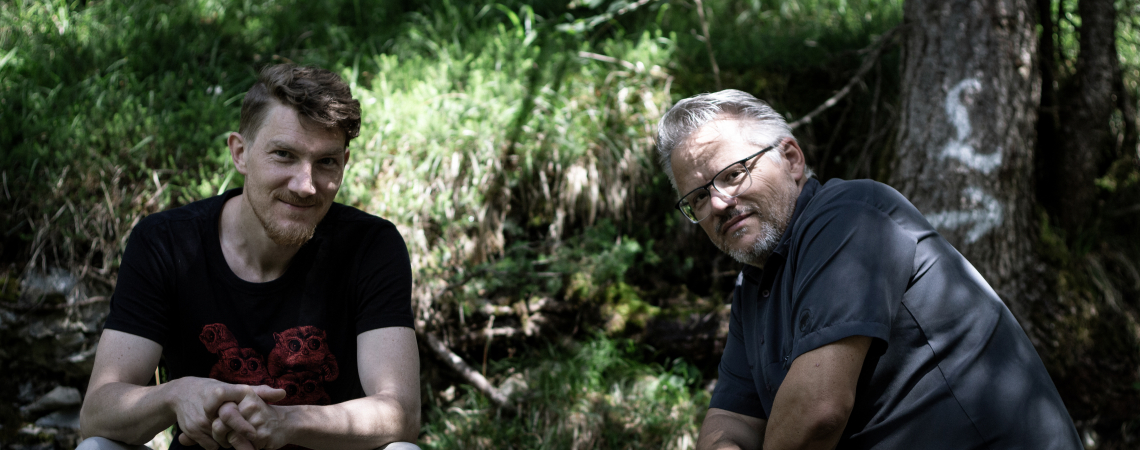 Zwei Männer sitzen im Wald, von Sonnenlicht durch Bäume beleuchtet.