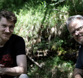 Zwei Männer sitzen im Wald, von Sonnenlicht durch Bäume beleuchtet.