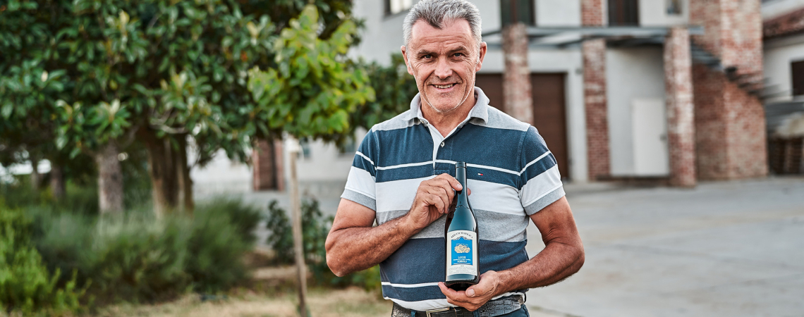 Mann hält Weinflasche im Freien, lächelnd vor Backsteingebäude und grüner Kulisse.
