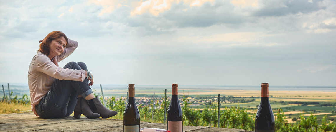 Frau am Weinhang mit drei Weinflaschen und Panoramablick über Landschaft.