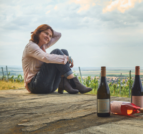 Frau am Weinhang mit drei Weinflaschen und Panoramablick über Landschaft.