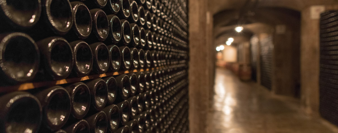 Weinkeller mit dicht gestapelten Flaschen entlang einer beleuchteten, rustikalen Steinwand.