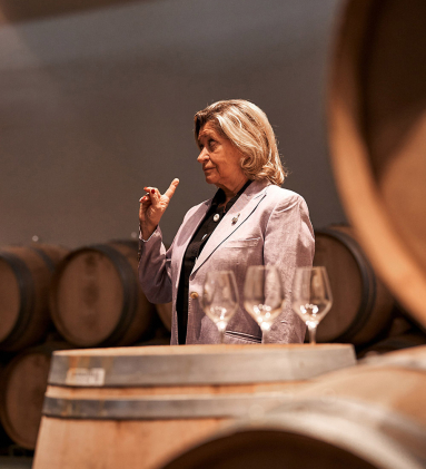 Frau gestikuliert vor Weinfässern, mit Gläsern auf einem Fass im Vordergrund.